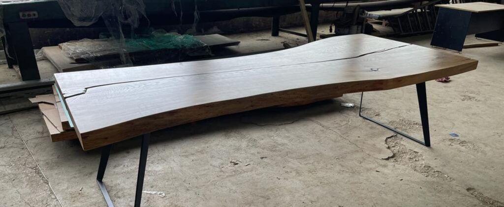 Live workshop shot of the Ragusa dining table: a large live-edge oak plank atop black steel legs