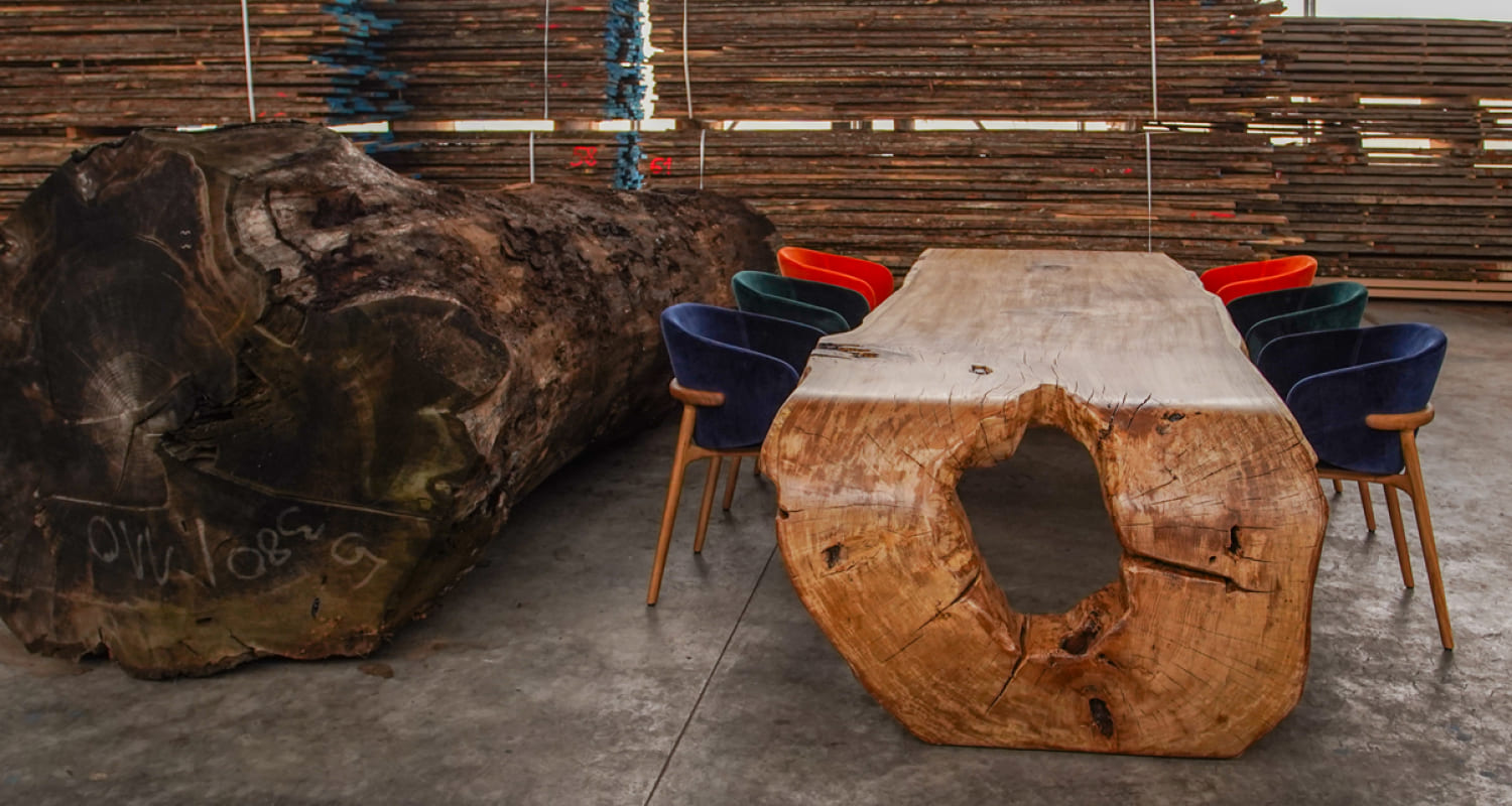 Ragusa solid oak table with sculpted log base placed next to raw oak trunk in workshop
